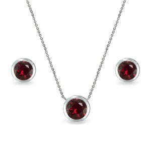 May include: A jewelry set featuring a necklace and matching stud earrings. The necklace has a silver chain with a large, round, deep red gemstone pendant. The earrings also have matching red gemstones set in silver bezels. The set is displayed on a white background.