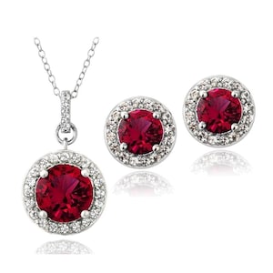 May include: A jewelry set featuring a pendant necklace and matching stud earrings. The necklace has a round, deep red gemstone pendant surrounded by a halo of small, clear stones. The earrings mirror the pendant design. The set is silver-toned.