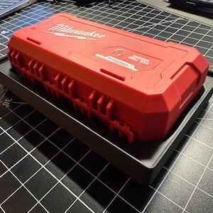 May include: A red Milwaukee tool case with the words "Milwaukee" and "Hammer Drill Bits" printed on the top. The case is sitting on a black tray. The background is a black surface with a grid pattern.
