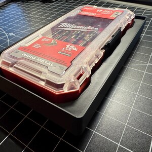 May include: A clear plastic case containing drill bits, labeled "Milwaukee" and "Hard Metal." The case is red and clear, with a black handle and is set in a black tray. The case has a red label with white text.