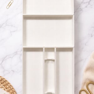 Puede incluir: Un conjunto de tres bandejas de almacenamiento rectangulares blancas, ideales para organizar objetos pequeños. Las bandejas están dispuestas verticalmente sobre una superficie de mármol, con artículos de oficina y elementos decorativos. Las bandejas están vacías.
