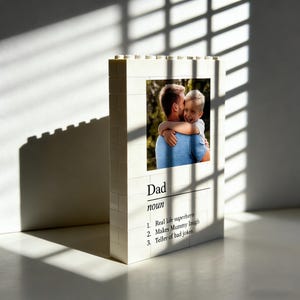 Puede incluir: Un marco de fotos blanco en forma de bloque con una foto de un hombre abrazando a un niño. El marco tiene la palabra "Papá" y una definición, que incluye "Superhéroe de la vida real" y "Cuentacuentos de chistes malos."