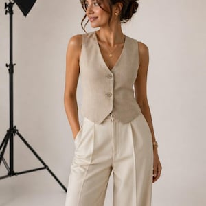 Puede incluir: Un chaleco de lino beige con tres botones y pantalones de pernera ancha y talle alto a juego. El conjunto es de color neutro y parece estar hecho de una tela ligera. El chaleco tiene cuello en V y los pantalones tienen pliegues.