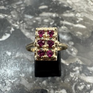 May include: A gold ring featuring a rectangular head with a textured surface and six round, deep pink gemstones arranged in two vertical columns of three. The band is thin and smooth, connecting to the sides of the rectangular setting.