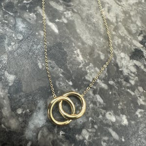 May include: A gold-toned necklace featuring a delicate chain and a pendant composed of two interlocking smooth circular rings. The metal has a polished, reflective finish, and the design consists of two simple, circular loops connected together.