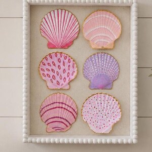 May include: A rectangular white beaded frame containing six decorative seashells arranged in two columns. The shells feature various hand-painted patterns in shades of pink, purple, and white, with gold-painted edges, set against a neutral background.