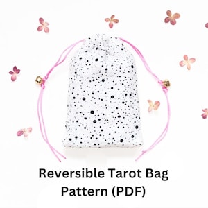 Puede incluir: Una bolsa de tarot reversible blanca con un patrón moteado negro. La bolsa tiene un cierre de cordón rosa con dijes de campana dorados. El texto "Reversible Tarot Bag Pattern (PDF)" se muestra debajo.