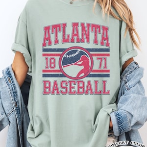 May include: A light green t-shirt with the words "ATLANTA BASEBALL" in red and blue lettering. The design includes a baseball player silhouette and the numbers "18" and "71". The shirt is labeled "COMFORT COLOR Bay".