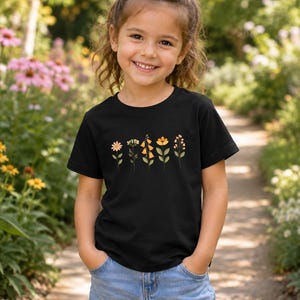May include: A black short-sleeved t-shirt featuring a graphic print of five distinct wildflowers in shades of yellow, orange, and green. The shirt is paired with light blue denim shorts that have a cuffed hem and visible front pockets.