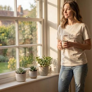 K&ouml;nnte beinhalten: Eine Frau tr&auml;gt ein cremefarbenes T-Shirt mit dem Text "Coffee & Sunshine" und einer Kaffeetasse, die eine Tasse h&auml;lt. Das Bild zeigt auch ein Fenster mit drei Topf-Sukkulenten auf der Fensterbank.