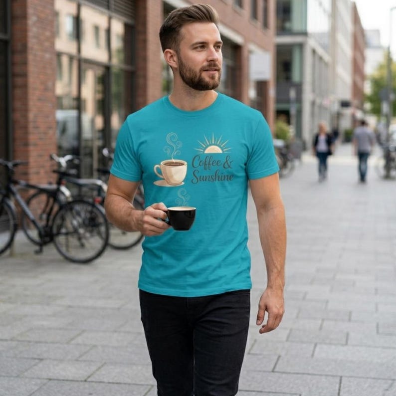 K&ouml;nnte beinhalten: T&uuml;rkisfarbenes T-Shirt mit einer Kaffeetasse und einem Sonnensymbol sowie dem Text "Coffee & Sunshine". Das kurz&auml;rmelige Shirt ist aus weichem Material. Die Person im Bild h&auml;lt eine schwarze Kaffeetasse.