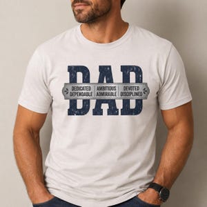 以下が含まれることがあります： ライトグレーの半袖Tシャツ。中央には、かすれた質感の大きなダークブルーの文字で「DAD」とプリントされています。その上に重なるシルバーの横長バナーには、「DEDICATED」「AMBITIOUS」「DEVOTED」「DEPENDABLE」「ADMIRABLE」「DISCIPLINED」という単語が記されています。