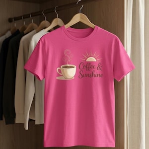 K&ouml;nnte beinhalten: Ein leuchtend pinkfarbenes T-Shirt mit einer Grafik einer Kaffeetasse, Dampf und einer Sonne mit dem Text "Coffee & Sunshine". Das Shirt h&auml;ngt an einem Holzb&uuml;gel in einem Schrank.