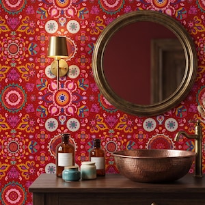 May include: A vibrant red wallpaper featuring a repeating intricate floral and geometric pattern in shades of pink, orange, blue, and white. The wall includes a round mirror with a dark frame, a gold wall sconce, and a copper sink on a wooden vanity.