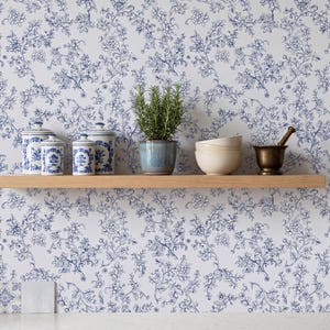 May include: A light-colored wooden shelf mounted against a wall covered in white wallpaper with a blue floral pattern. On the shelf are two blue and white patterned canisters, a green plant in a blue pot, two stacked cream bowls, and a brass mortar.