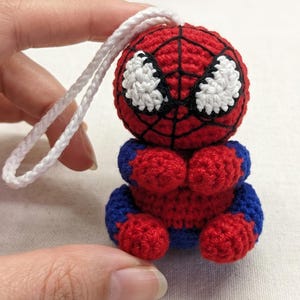 May include: A small, round, crocheted figure resembling a superhero with a red and blue body, black web-like stitching, and large white eyes. The figure features a white braided cord loop attached to the top for hanging as an ornament.