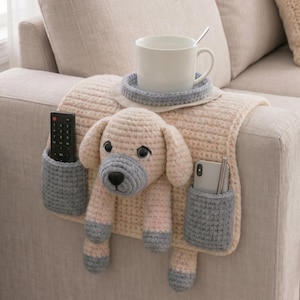 May include: A crocheted sofa armrest featuring a dog design. The beige armrest has two gray pockets for a remote and phone, and a cup holder with a blue coaster. A white mug with a spoon rests on the coaster.