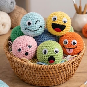 Puede incluir: Una colección de bolas de hilo tejidas a ganchillo con ojos móviles y expresiones faciales bordadas, incluyendo caras guiñando, sonriendo, sorprendidas y sacando la lengua, dispuestas dentro de una cesta de mimbre con varios colores de hilo.
