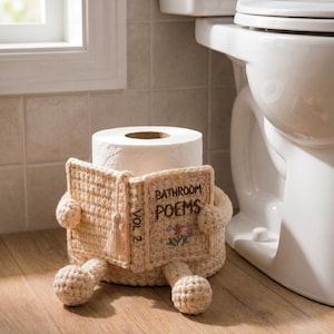May include: A cream-colored crochet toilet paper roll cover shaped like a seated figure holding an open book. The book cover features the embroidered text "BATHROOM POEMS" and "VOL 2" with a small floral design and a decorative tassel.