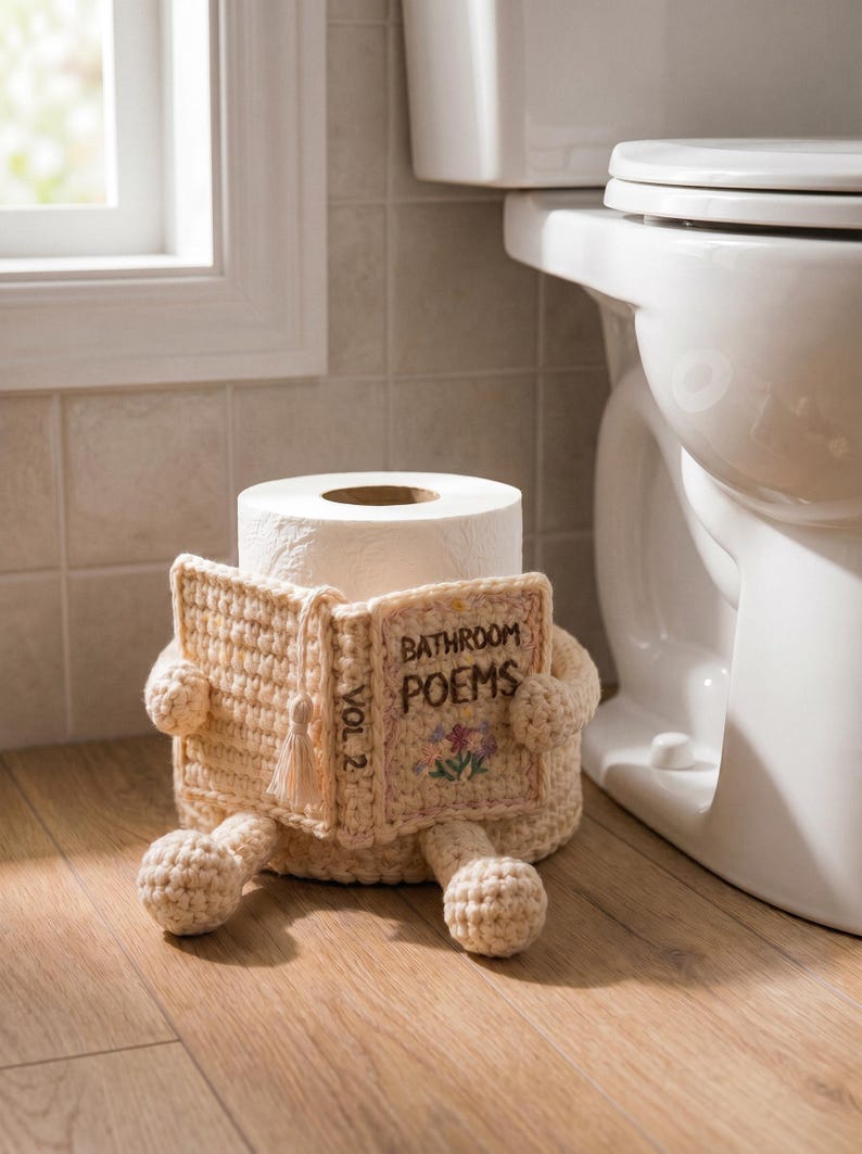 May include: A cream-colored crochet toilet paper roll cover shaped like a seated figure holding an open book. The book cover features the embroidered text "BATHROOM POEMS" and "VOL 2" with a small floral design and a decorative tassel.