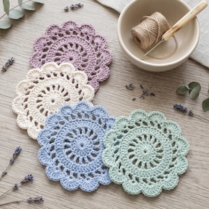May include: Four handmade crochet coasters in pastel colors: lavender, cream, light blue, and mint green. A small bowl with twine and a crochet hook are also present. The coasters are circular with a scalloped edge, perfect for home decor.