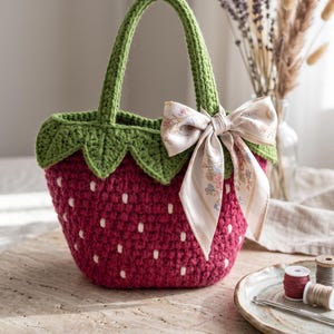 May include: A handmade crochet handbag shaped like a strawberry, featuring a deep red body with small white embroidered seeds, a green leafy top border, a green handle, and a light-colored silk ribbon tied in a bow on the side of the bag.