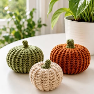 May include: Three crocheted pumpkins in olive green, cream, and burnt orange. Each pumpkin features a ribbed texture and a small, solid green crocheted stem at the top. The pumpkins are arranged in a cluster, showcasing their distinct colors.