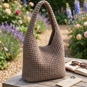May include: A taupe-coloured hobo-style bag crafted with a dense, textured crochet stitch pattern. The bag features a single integrated shoulder strap that forms a continuous loop with the main body, creating a structured yet flexible silhouette.