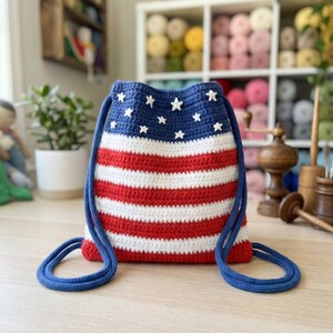 Puede incluir: Una mochila de crochet con cierre de cord&oacute;n que presenta un patr&oacute;n inspirado en la bandera estadounidense, con una secci&oacute;n superior azul oscuro adornada con peque&ntilde;as estrellas blancas y rayas horizontales rojas y blancas, con correas de cuerda azul.