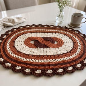 Op de afbeelding: Een ovaal gehaakt placemat met concentrische ringen in tinten donkerbruin, roestoranje en crème. Het midden toont een in elkaar grijpend oneindigheidslusontwerp, omringd door gestructureerde steken en een geschulpte, kanten rand.