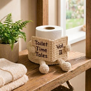 May include: A cream-colored crocheted toilet paper roll cover shaped like a book with small arms and legs. The front of the cover features the embroidered dark brown text Toilet Time Tales Vol. II. The cover wraps around a standard white toilet paper roll.