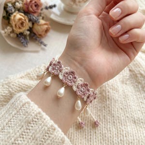 May include: A crochet bracelet featuring pink and white floral designs with dangling faux pearls. The bracelet is worn on a wrist, with a cream-colored sweater in the background. A bouquet of dried flowers and a teacup are also visible.