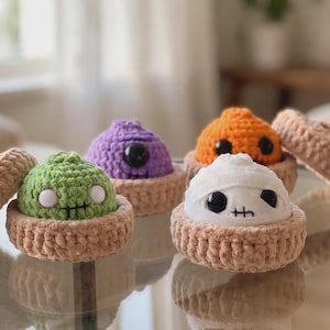 May include: Four small, round, crocheted figures nestled in tan, bowl-shaped bases. The figures include a green one with white eyes, a purple one with a single black eye, an orange one with black eyes, and a white mummy-like figure with black eyes.