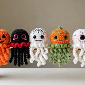 May include: Five crocheted plush toys shaped like jellyfish with Halloween themes. The figures include an orange jack-o'-lantern, a black creature with red eyes, a white ghost with a spiderweb, an orange pumpkin with green tentacles, and a white eyeball.