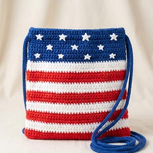 Puede incluir: Un bolso bandolera rectangular de ganchillo con un dise&ntilde;o patri&oacute;tico. La secci&oacute;n superior es azul oscuro con peque&ntilde;as estrellas blancas tejidas, mientras que la parte inferior presenta rayas horizontales rojas y blancas alternas. Incluye una correa azul.