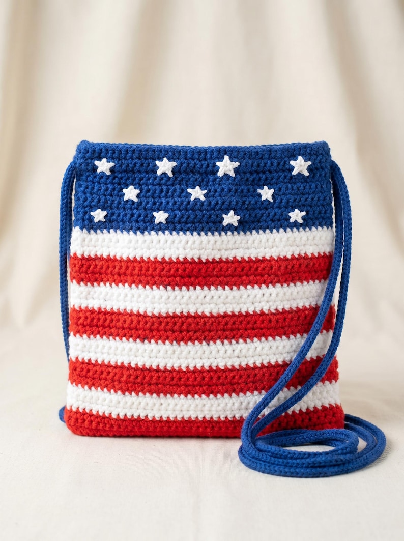 Puede incluir: Un bolso bandolera rectangular de ganchillo con un dise&ntilde;o patri&oacute;tico. La secci&oacute;n superior es azul oscuro con peque&ntilde;as estrellas blancas tejidas, mientras que la parte inferior presenta rayas horizontales rojas y blancas alternas. Incluye una correa azul.