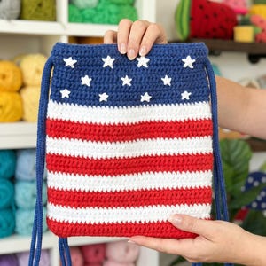 Puede incluir: Una bolsa de ganchillo rectangular con cord&oacute;n que presenta un dise&ntilde;o patri&oacute;tico con una secci&oacute;n superior azul oscuro adornada con estrellas blancas y una secci&oacute;n inferior de rayas horizontales rojas y blancas, con correas de cord&oacute;n azul oscuro.