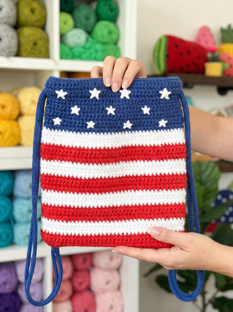 Puede incluir: Una bolsa de ganchillo rectangular con cord&oacute;n que presenta un dise&ntilde;o patri&oacute;tico con una secci&oacute;n superior azul oscuro adornada con estrellas blancas y una secci&oacute;n inferior de rayas horizontales rojas y blancas, con correas de cord&oacute;n azul oscuro.