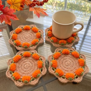 May include: Four round, beige crochet coasters featuring a scalloped edge and a border of small, bright orange pumpkin-shaped yarn embellishments. One coaster supports a plain, off-white ceramic mug. The design is consistent across all four items.