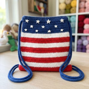 Puede incluir: Una mochila de crochet con cord&oacute;n que presenta un patr&oacute;n de bandera en rojo, blanco y azul. La secci&oacute;n superior es azul oscuro con peque&ntilde;as estrellas blancas tejidas, mientras que la inferior tiene rayas horizontales rojas y blancas.