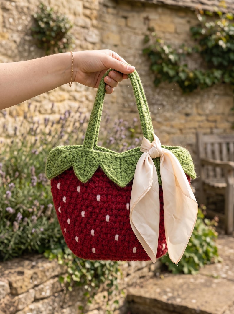 May include: A handmade crochet tote bag shaped like a strawberry, featuring a deep red body with small white embroidered seeds, a green leafy top border, two green handles, and a light cream-colored silk ribbon tied in a bow on the side.