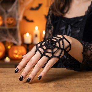 Peut inclure: Un accessoire de main en crochet noir présentant un motif de toile d'araignée qui couvre le dos de la main et se connecte à une boucle de doigt. Le design complexe en fil crée une structure géométrique qui repose sur les articulations.
