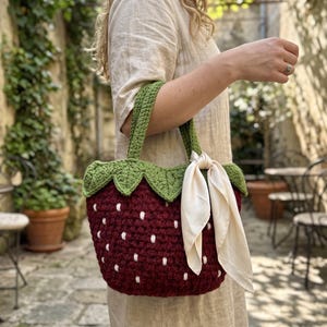 May include: A dark red crochet handbag shaped like a strawberry with white embroidered seeds, a green leafy top border, two green crochet handles, and a cream-colored silk ribbon tied to one handle, creating a textured and functional accessory.