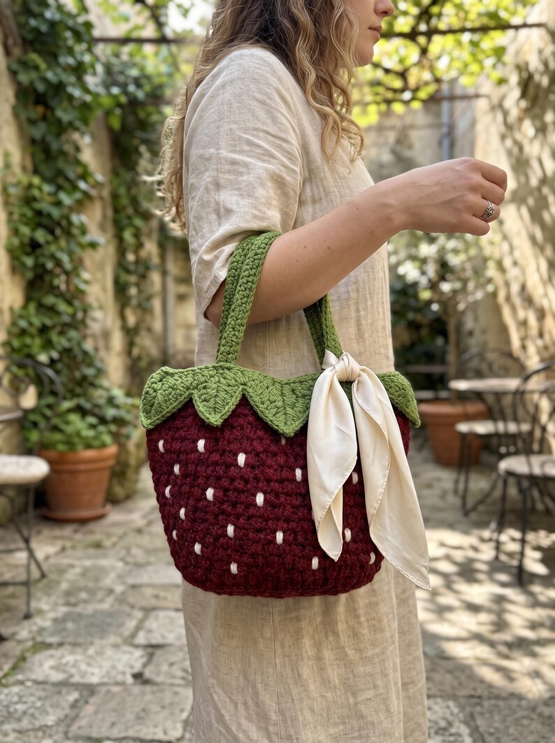 May include: A dark red crochet handbag shaped like a strawberry with white embroidered seeds, a green leafy top border, two green crochet handles, and a cream-colored silk ribbon tied to one handle, creating a textured and functional accessory.