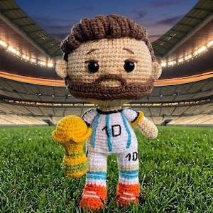 Puede incluir: Una muñeca de ganchillo con cabello castaño y barba, que viste una camiseta blanca con rayas azul claro y el número 10 en negro. La muñeca sostiene un trofeo amarillo y verde. La figura tiene ojos negros y usa calzado naranja y blanco.