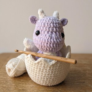 May include: A crocheted lavender and white dragon sits in a crocheted eggshell with a wooden crochet hook. The dragon has black eyes and white horns. A ball of white yarn rests next to the egg.