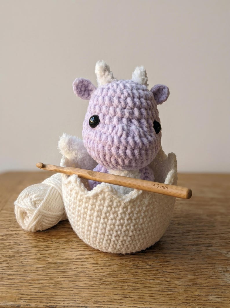 May include: A crocheted lavender and white dragon sits in a crocheted eggshell with a wooden crochet hook. The dragon has black eyes and white horns. A ball of white yarn rests next to the egg.