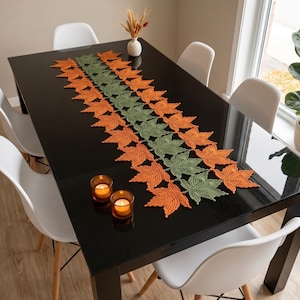 Puede incluir: Un camino de mesa largo y rectangular elaborado con hojas de arce de ganchillo en filas alternas de color naranja y verde salvia. El camino presenta un patrón repetitivo de formas de hojas individuales unidas para crear una pieza decorativa.