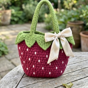 May include: A crochet handbag shaped like a strawberry, featuring a deep red body with small white embroidered dots, a green leafy top border, a single green handle, and a decorative cream-colored satin ribbon bow tied to the side of the handle.