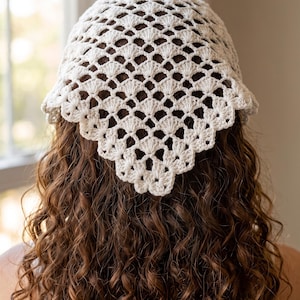 May include: A triangular head scarf made of off-white yarn featuring a delicate, openwork crochet pattern with repeating shell-like motifs and a scalloped edge. The accessory is designed to be worn over the hair, covering the crown area.